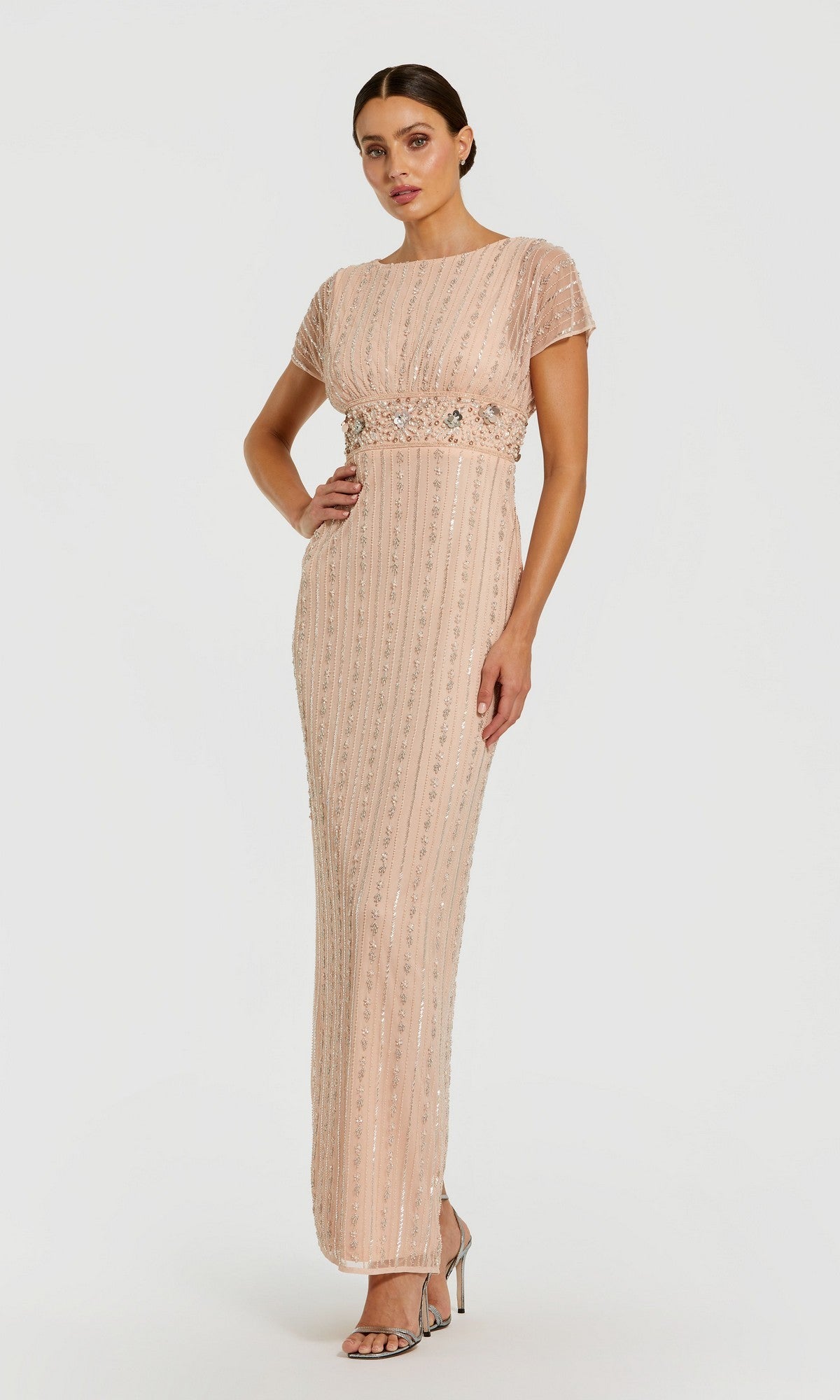 A woman stands against a plain white background wearing the Mac Duggal 94147 long formal dress in rose—a floor-length gown with short sleeves and beaded pearl embellishments—paired with silver high-heeled sandals.